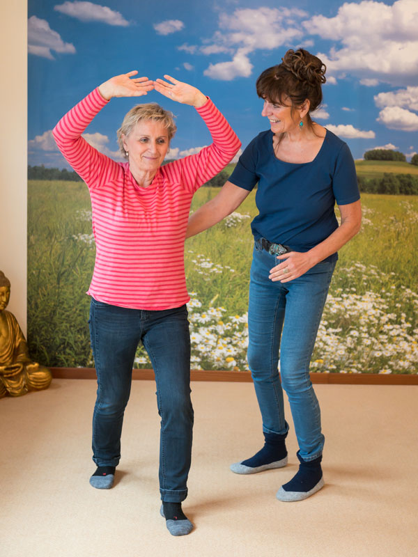 Petra Stolle zeigt einer Frau Qigong Übungen. Im Hintergrund an der Wand ist eine Wandtapete mit einer blühenden Frühlingswiese und blauem Himmel mit Schäfchenwolken.